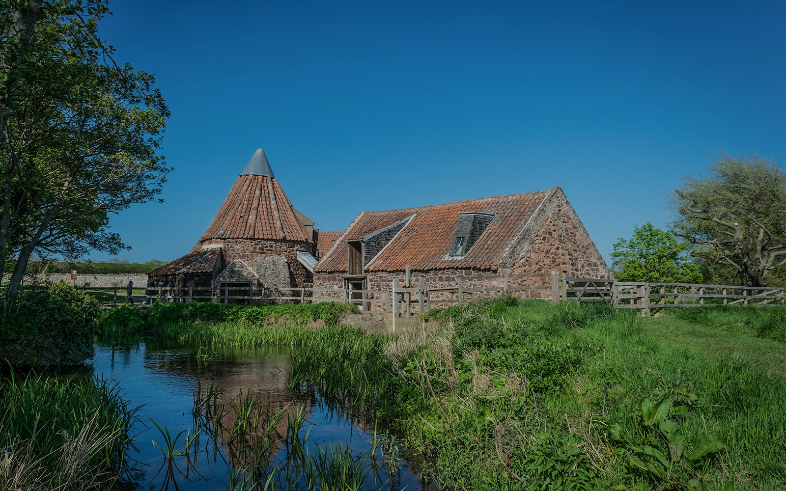 Mill building with red-tiled roof at Hobbiton Movie Set, surrounded by greenery and a stream.