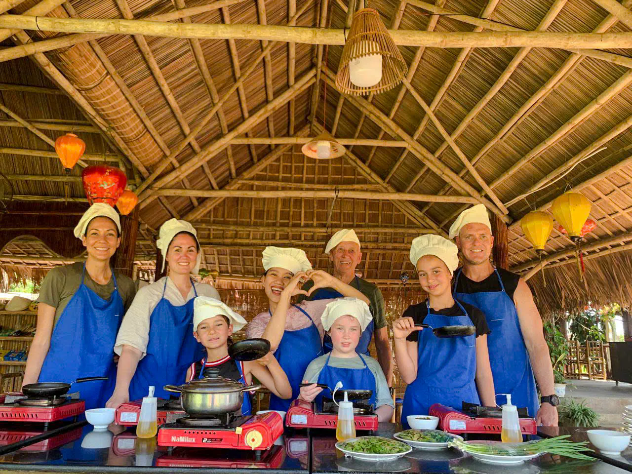 Group cooking class in Hoi An village, participants in aprons and chef hats.