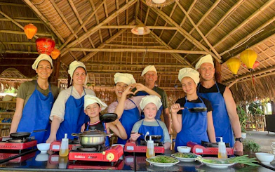Group cooking class in Hoi An village, participants in aprons and chef hats.