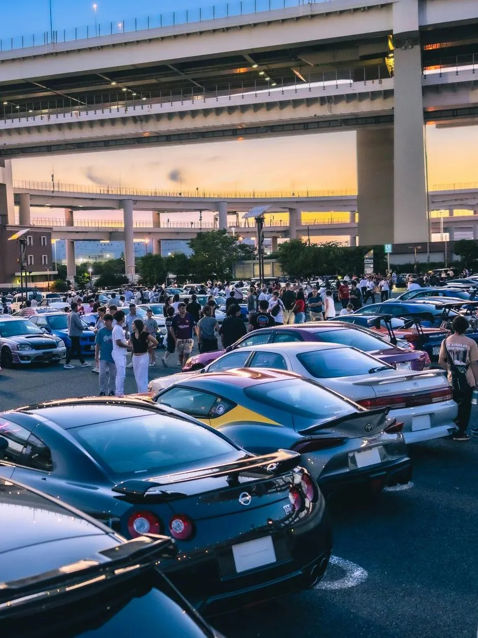 Car enthusiasts gather at Daikoku PA in Tokyo, showcasing a variety of vehicles under highway overpasses.