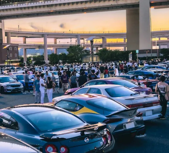 Car enthusiasts gather at Daikoku PA in Tokyo, showcasing a variety of vehicles under highway overpasses.