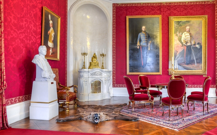 Interior of Royal Palace of Gödöllő with red walls, portraits, and ornate furniture.