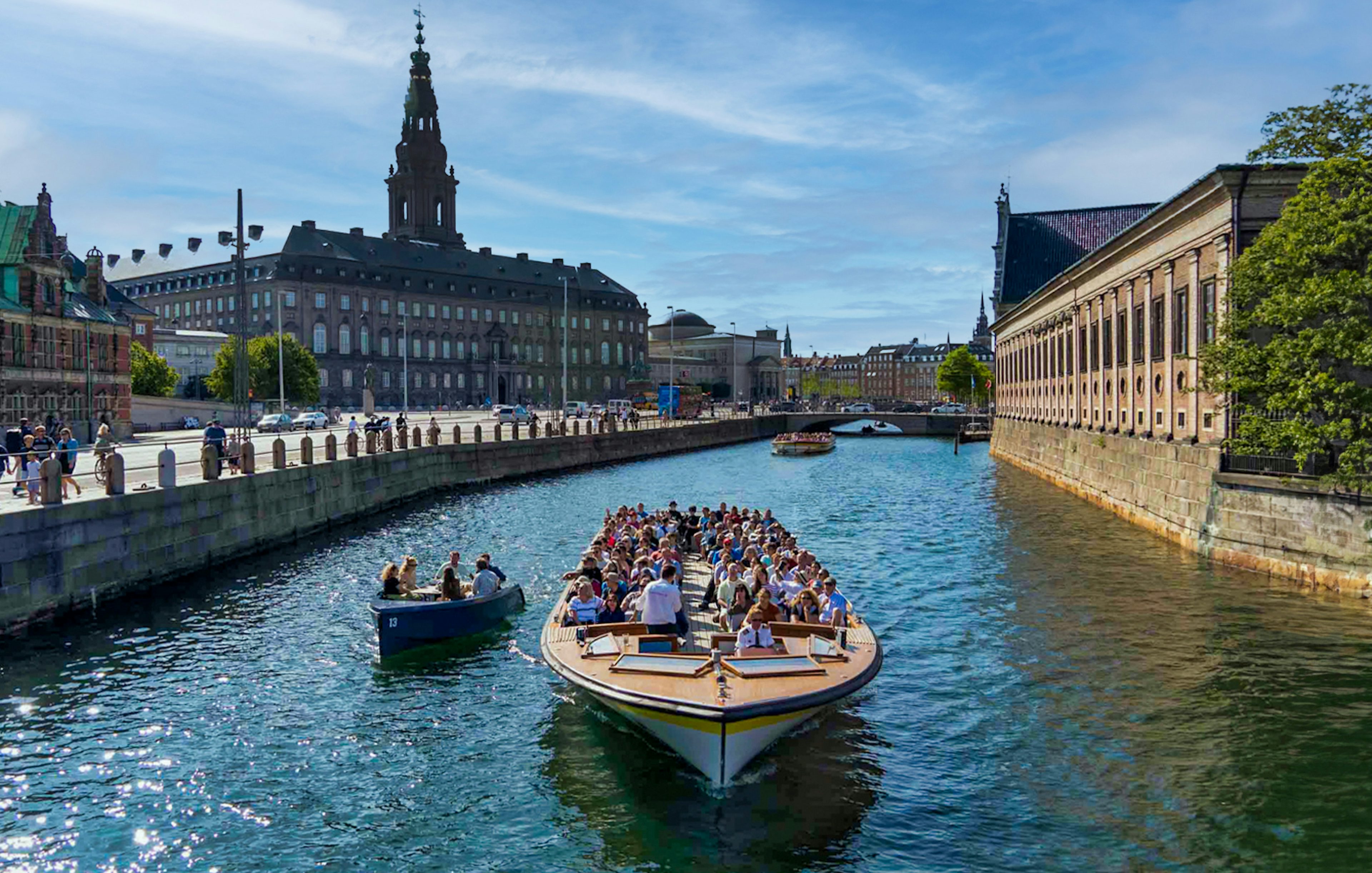 Canal cruise boat with tourists in Copenhagen, passing historic buildings and a bridge.