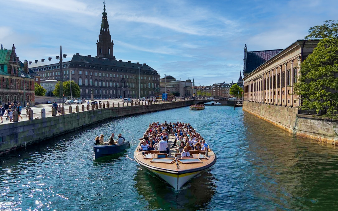 Canal cruise boat with tourists in Copenhagen, passing historic buildings and a bridge.