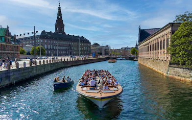Canal cruise boat with tourists in Copenhagen, passing historic buildings and a bridge.