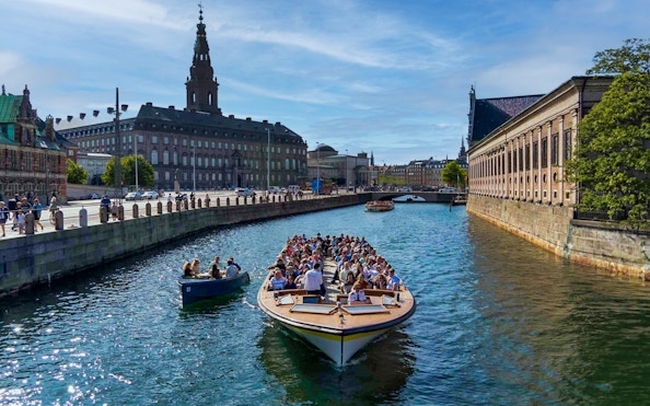 Canal cruise boat with tourists in Copenhagen, passing historic buildings and a bridge.
