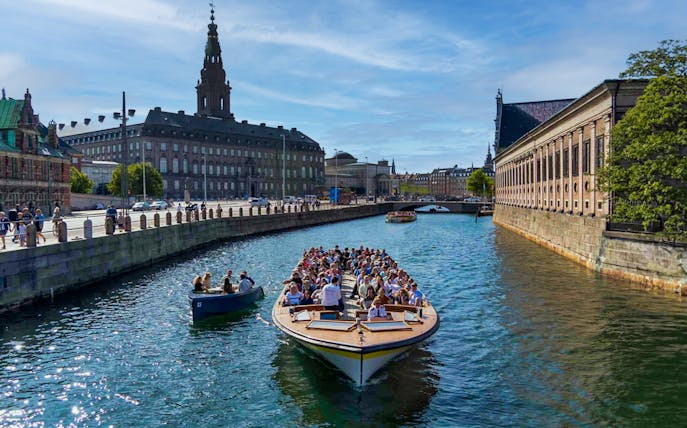 Canal cruise boat with tourists in Copenhagen, passing historic buildings and a bridge.