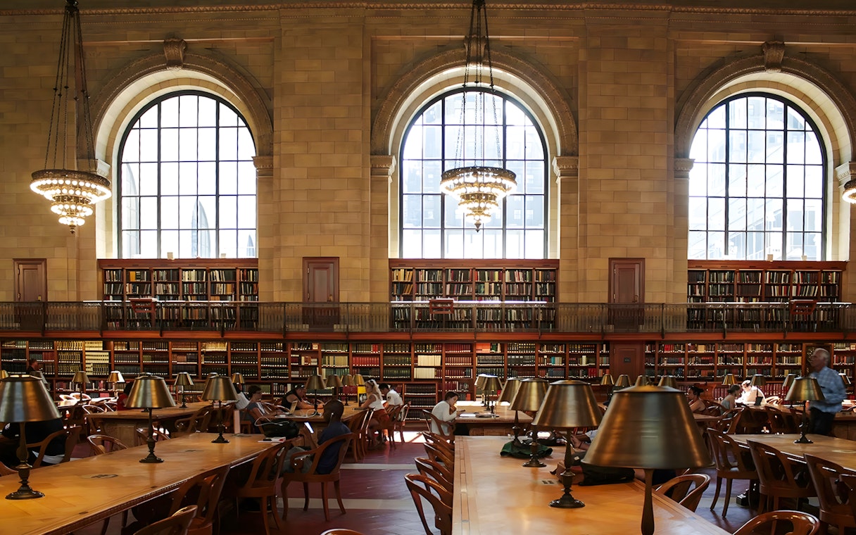 Interior reading room of the New York Public Library Stephen A. Schwarzman Building with large windows and chandeliers.