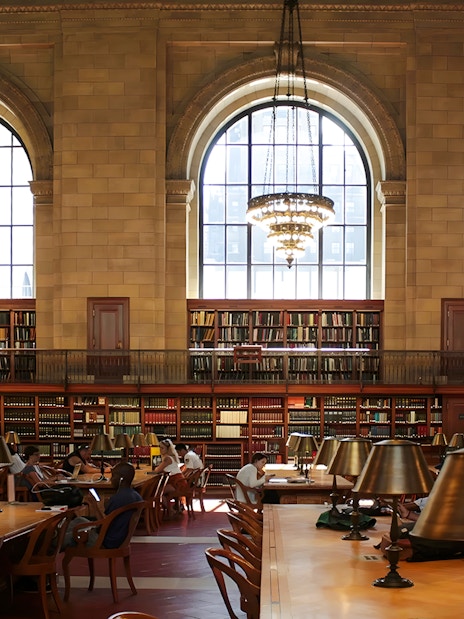 Interior reading room of the New York Public Library Stephen A. Schwarzman Building with large windows and chandeliers.