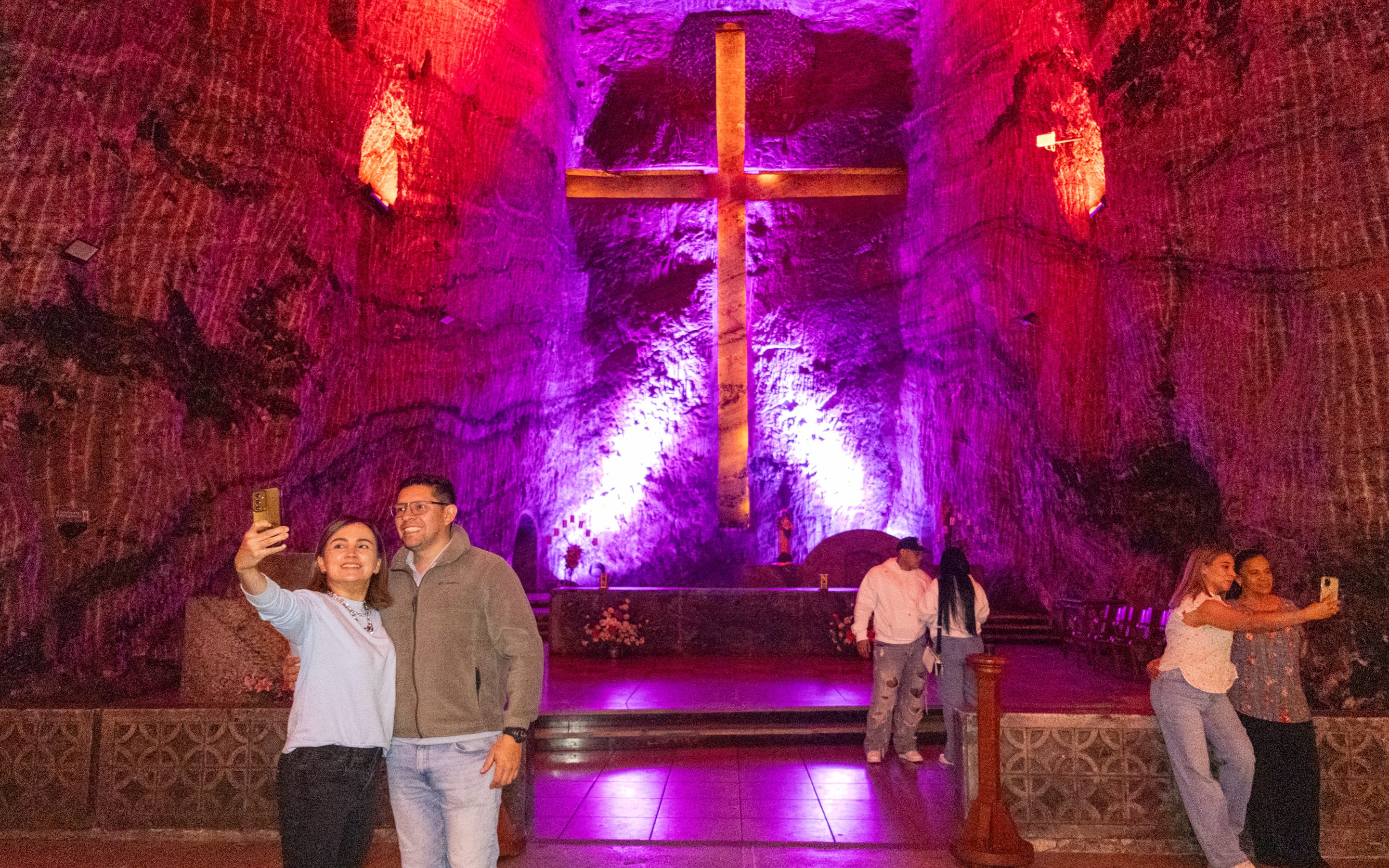 Tourists taking selfies inside Zipaquira Salt Cathedral with illuminated cross in background.