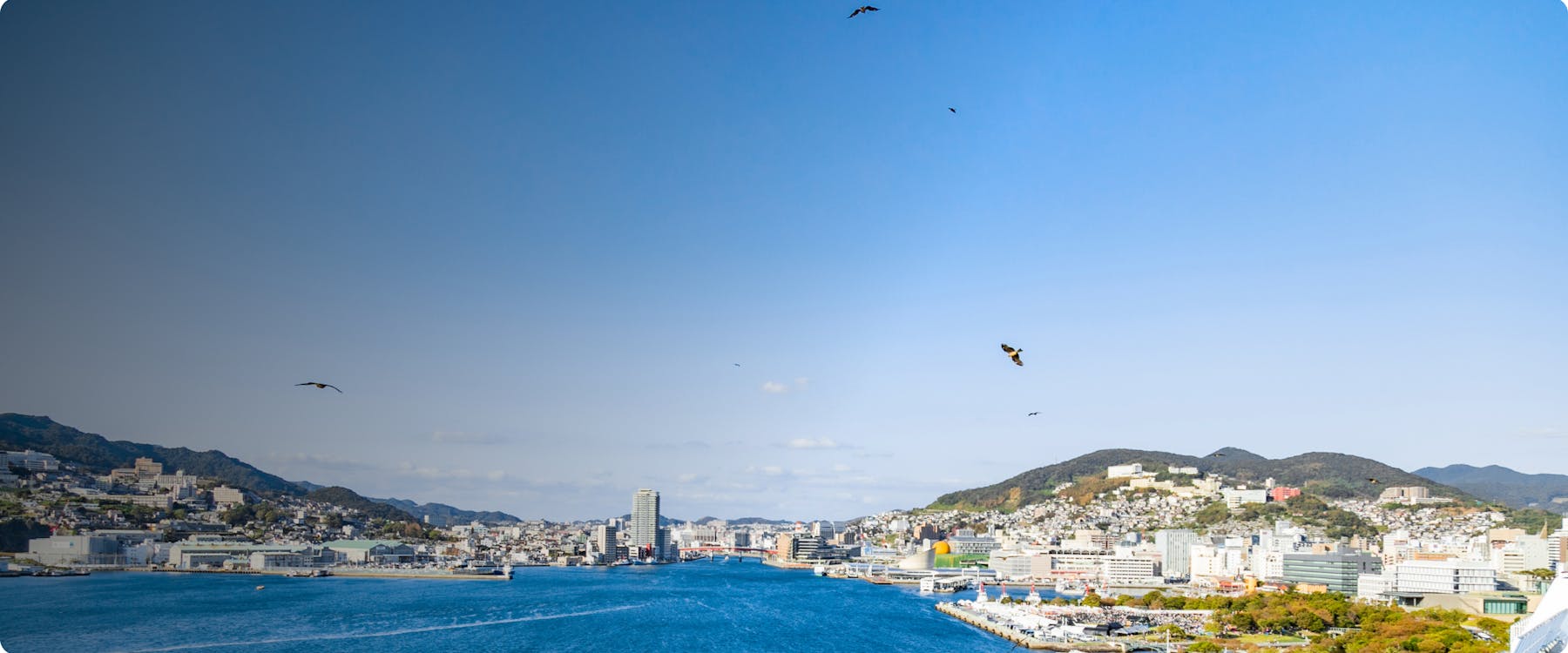 Nagasaki cityscape with harbor and surrounding hills under a clear blue sky.