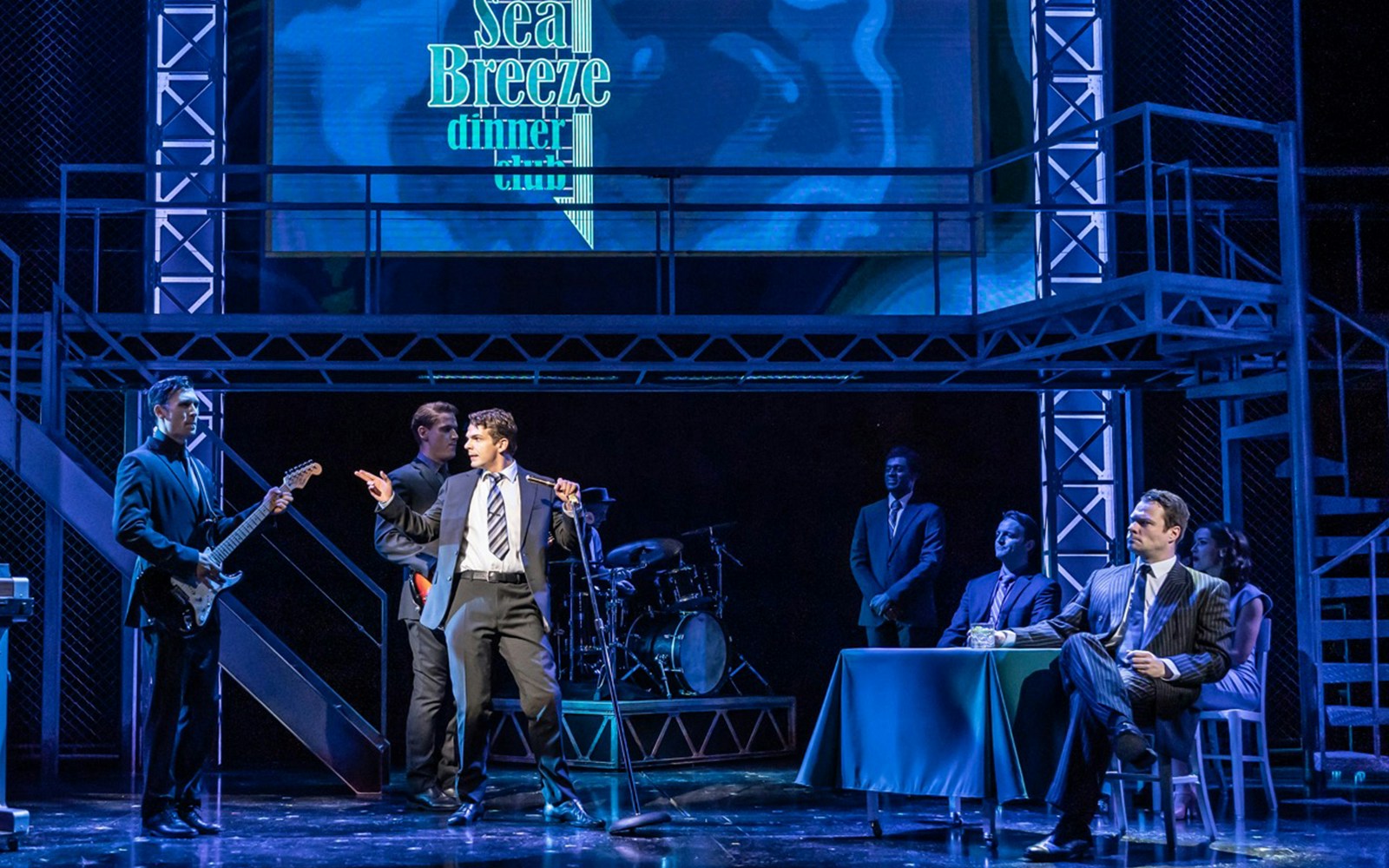 Stage performance of Jersey Boys with actors singing and playing instruments.