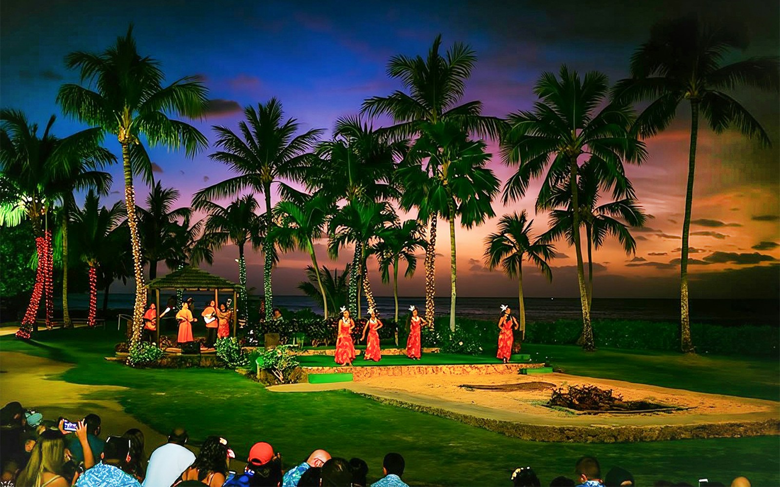 Dancers performing at Paradise Cove Luau, Hawaii, with palm trees and sunset in the background.