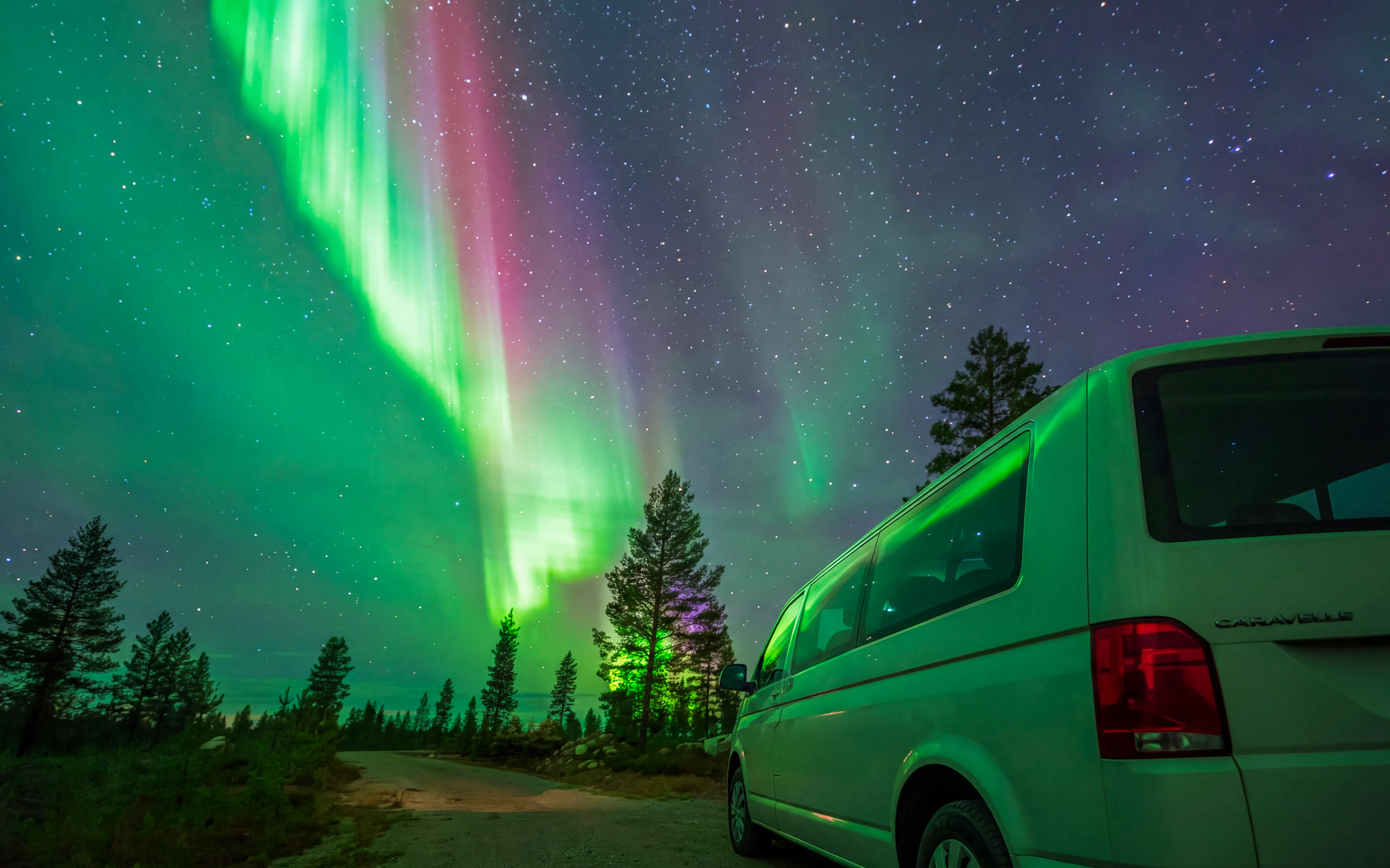 Van parked under vibrant Northern Lights in a forest setting.
