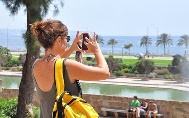 Woman taking photo near Cathedral of Palma with sea and palm trees in background.