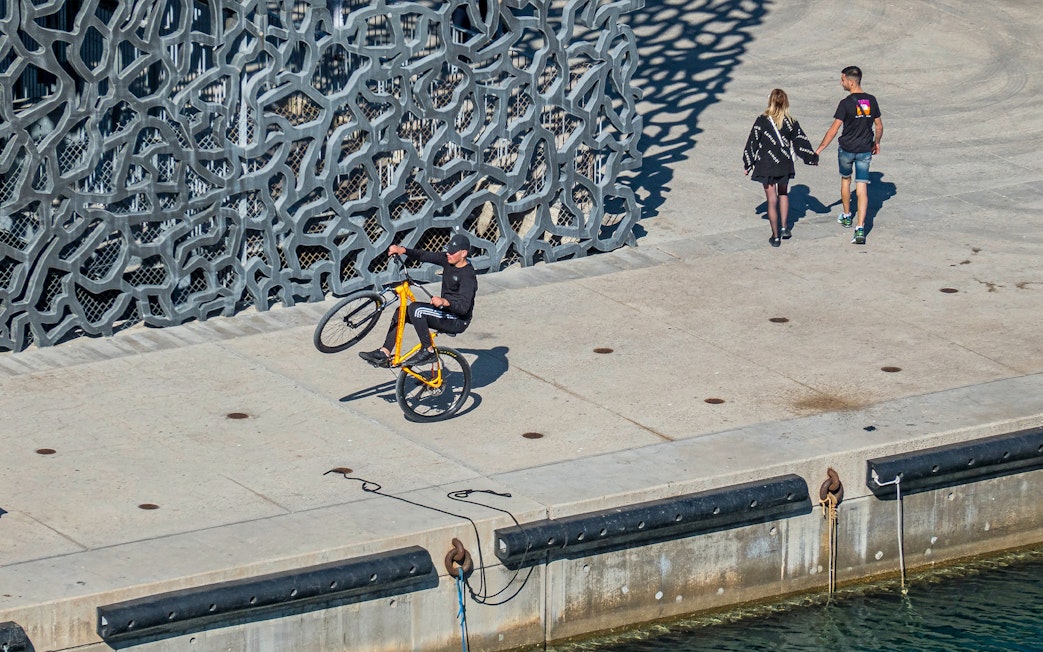 Cyclist performing a wheelie near Mucem's unique facade in Marseille.