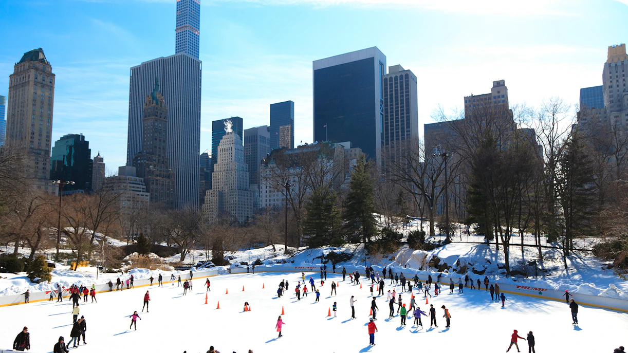 Ice skaters at Central Park Wollman Rink with New York City skyline in the background.