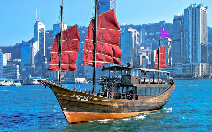 AquaLuna boat with red sails cruising Victoria Harbour, Hong Kong skyline in background.
