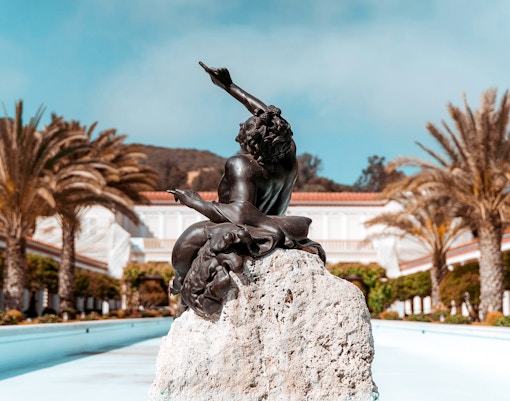 Statue in courtyard of Villa of the Papyri with palm trees and building in background.