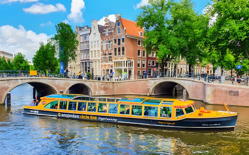 Amsterdam canal cruise boat passing under a bridge with historic buildings in the background.