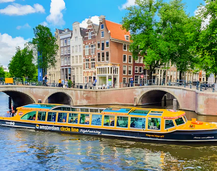 Amsterdam canal cruise boat passing under a bridge with historic buildings in the background.