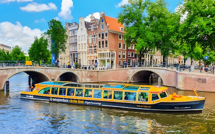 Amsterdam canal cruise boat passing under a bridge with historic buildings in the background.