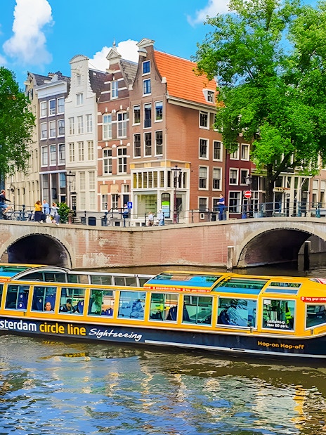 Amsterdam canal cruise boat passing under a bridge with historic buildings in the background.