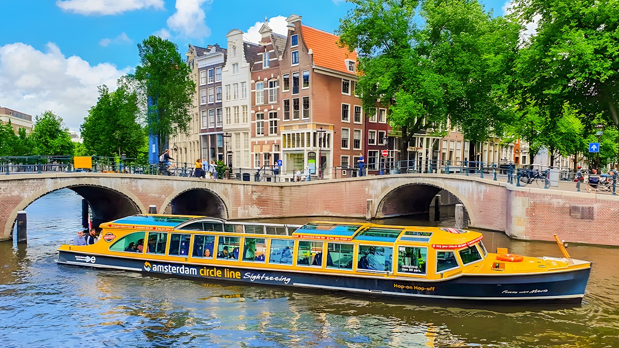 Amsterdam canal cruise boat passing under a bridge with historic buildings in the background.