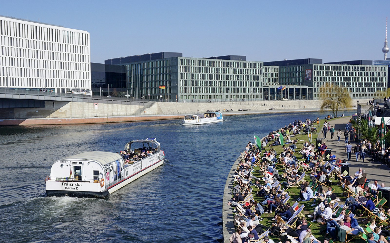 River cruise boat on Spree River in Berlin with passengers and riverside crowd.