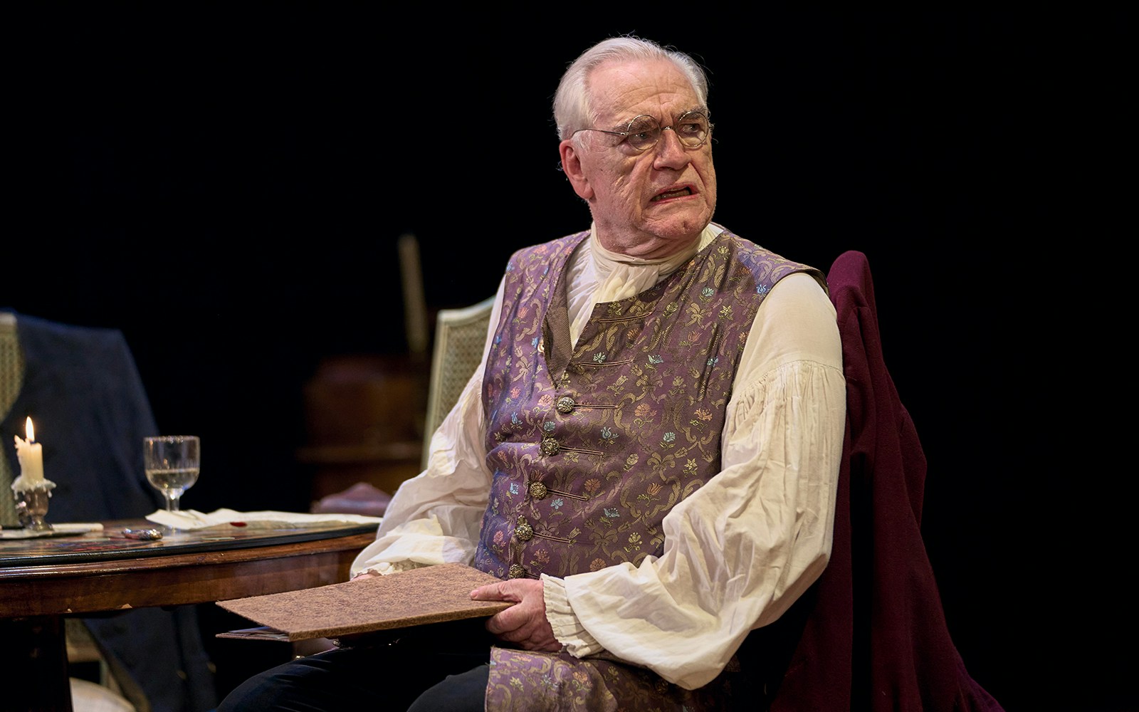 Elderly man in period costume sitting at a table with a candle and papers, performing in "The Score.