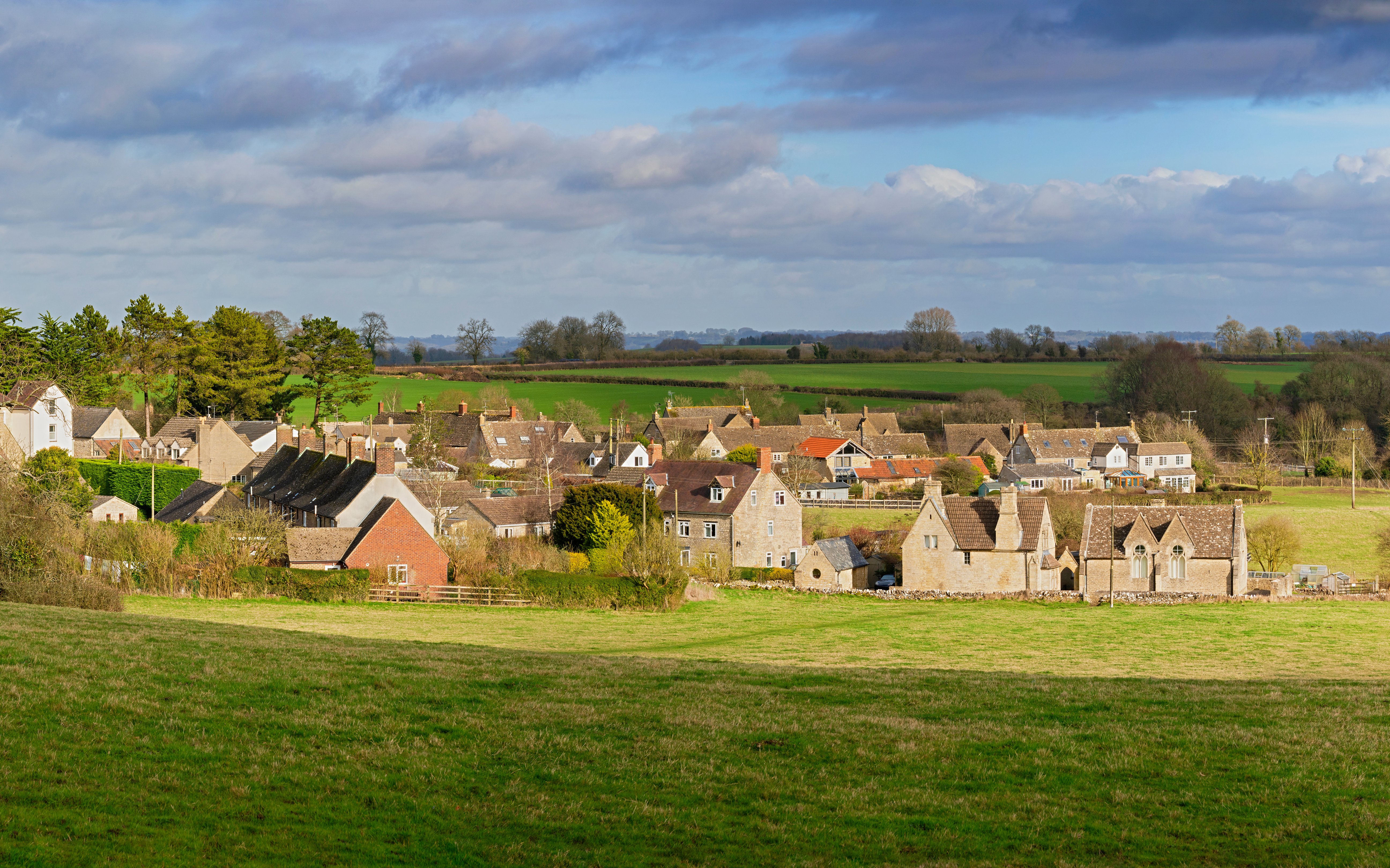 Cotswold village with stone cottages and green fields under a cloudy sky.