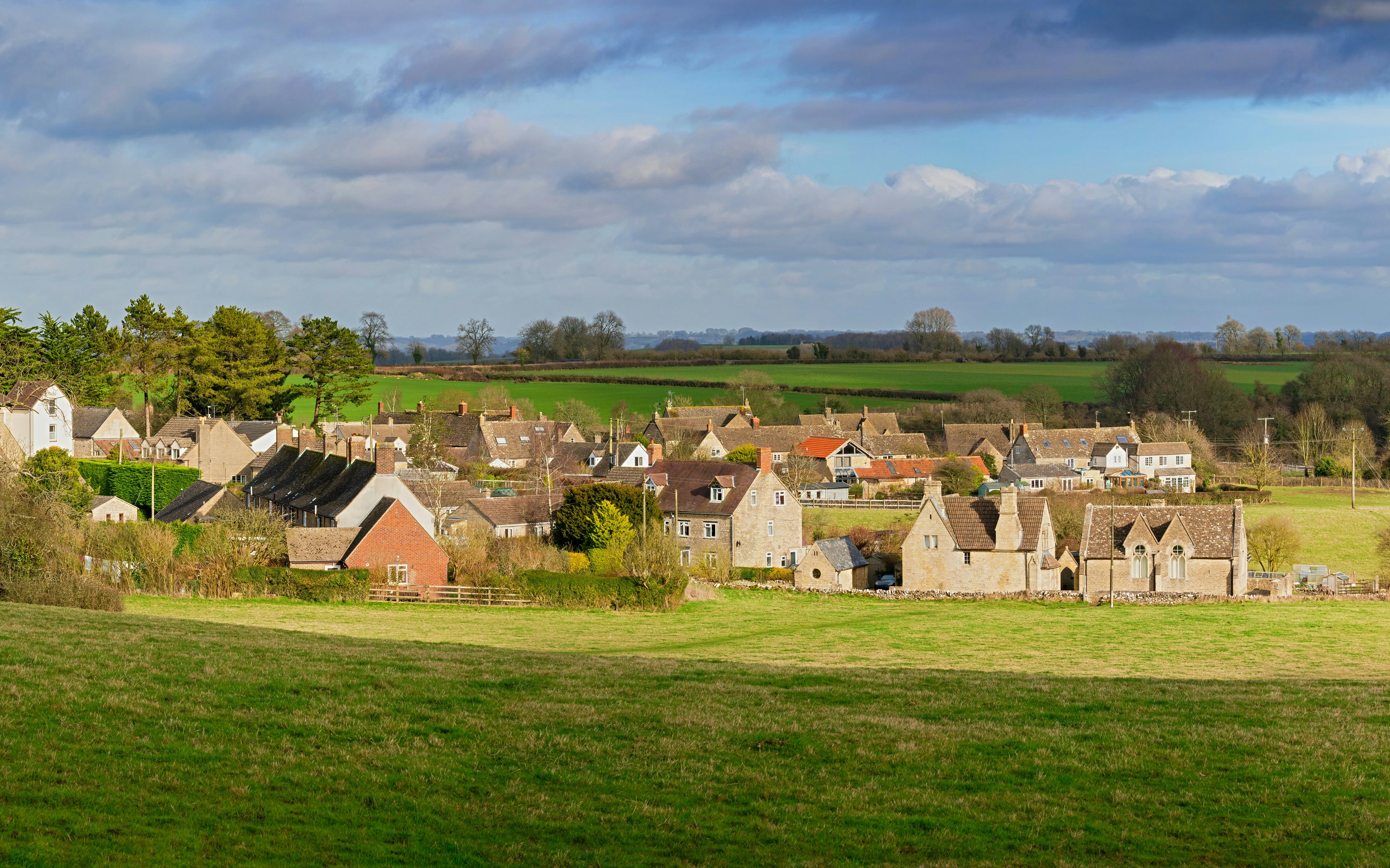 Cotswold village with stone cottages and green fields under a cloudy sky.