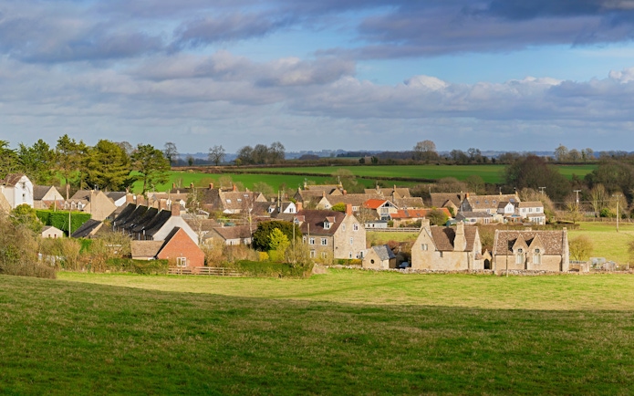 Cotswold village with stone cottages and green fields under a cloudy sky.