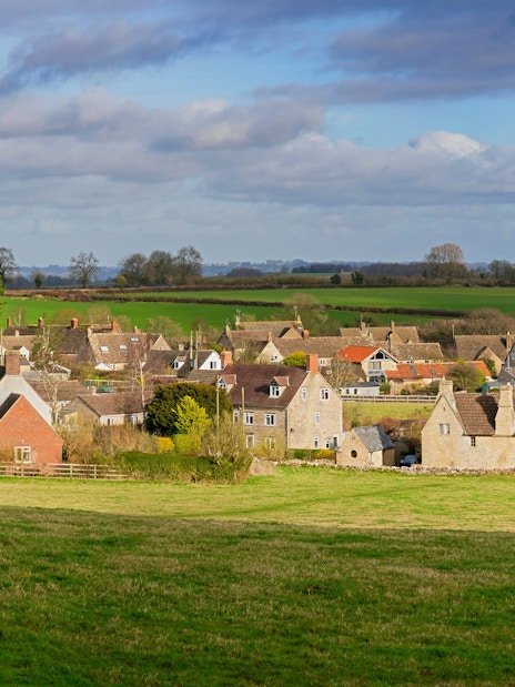 Cotswold village with stone cottages and green fields under a cloudy sky.