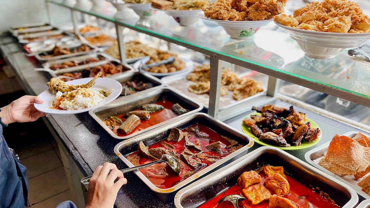 Padang food stall with various dishes and a person serving rice and fish.