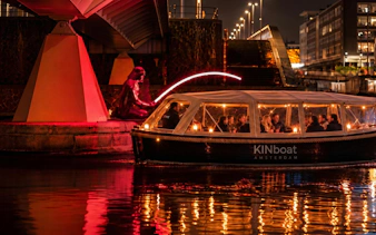 Canal boat tour during Amsterdam Light Festival with illuminated sculpture under a bridge.
