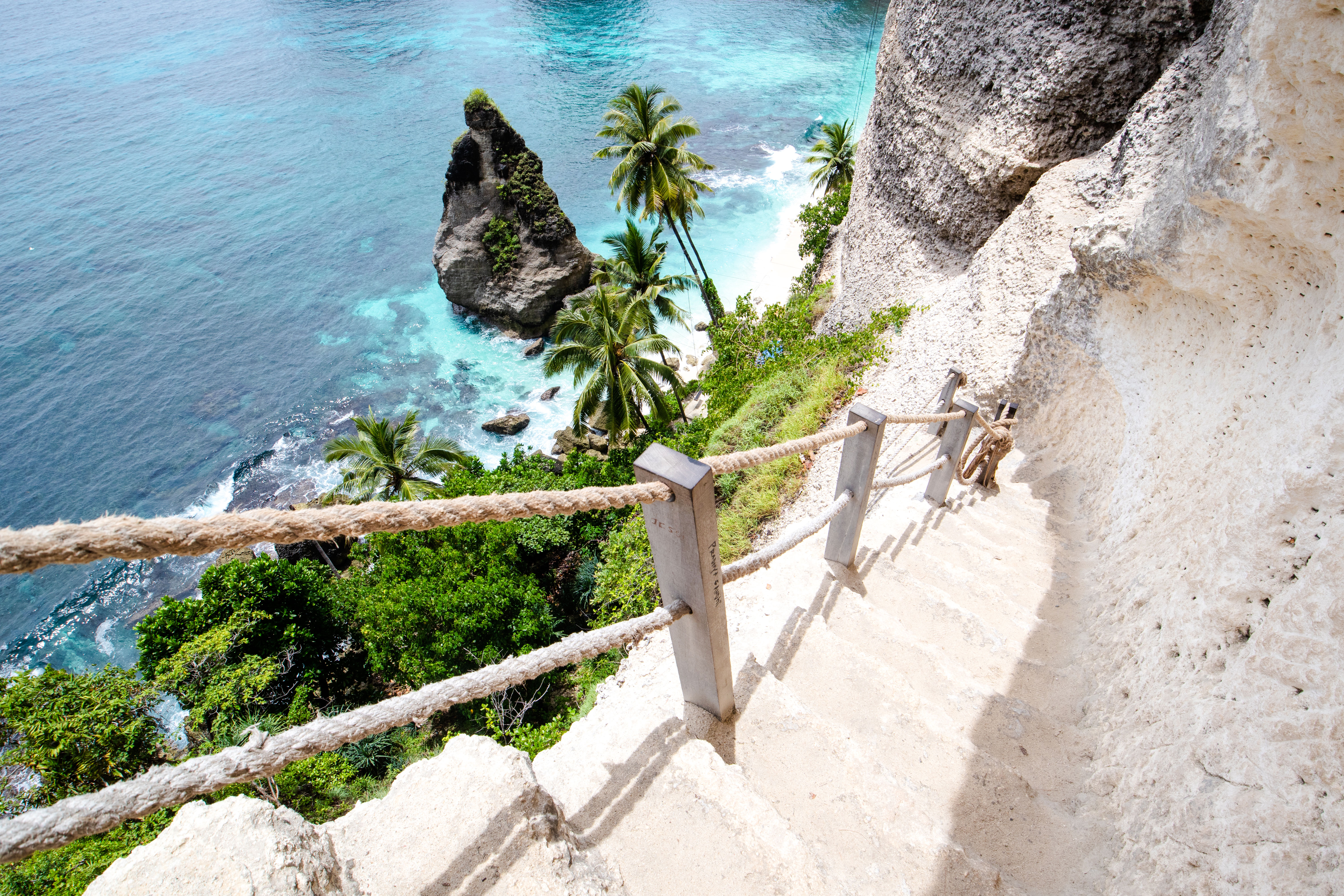 Steep stone stairs with rope railing leading down to a tropical beach in Bali.
