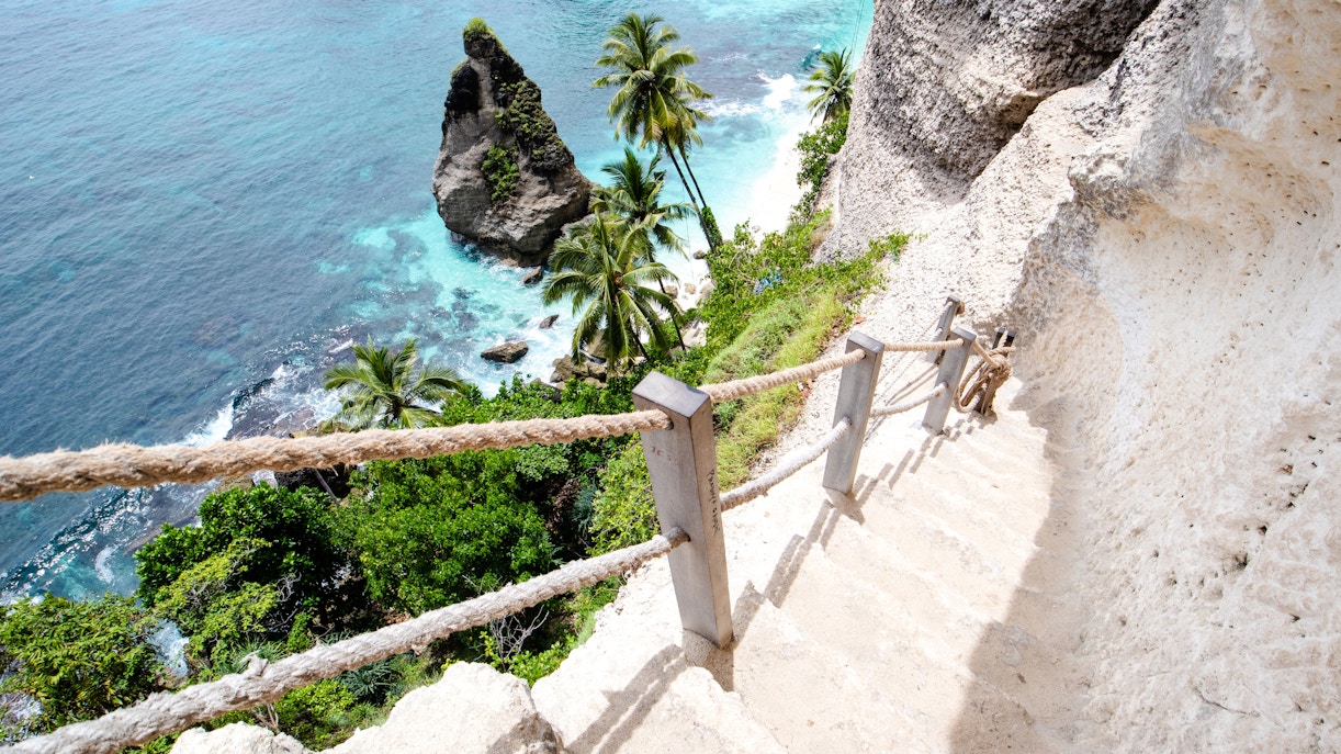 Steep stone stairs with rope railing leading down to a tropical beach in Bali.