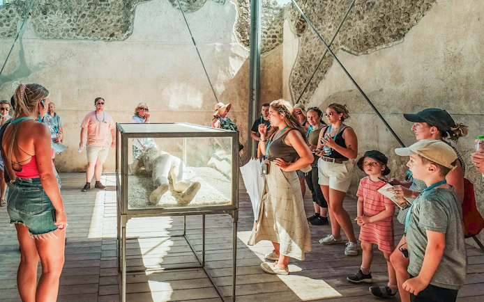 Tour group observing a plaster cast of a victim at Pompeii with a guide explaining.