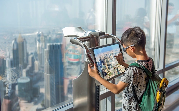Child using electronic telescope at Level 124, Burj Khalifa, viewing Dubai skyline.