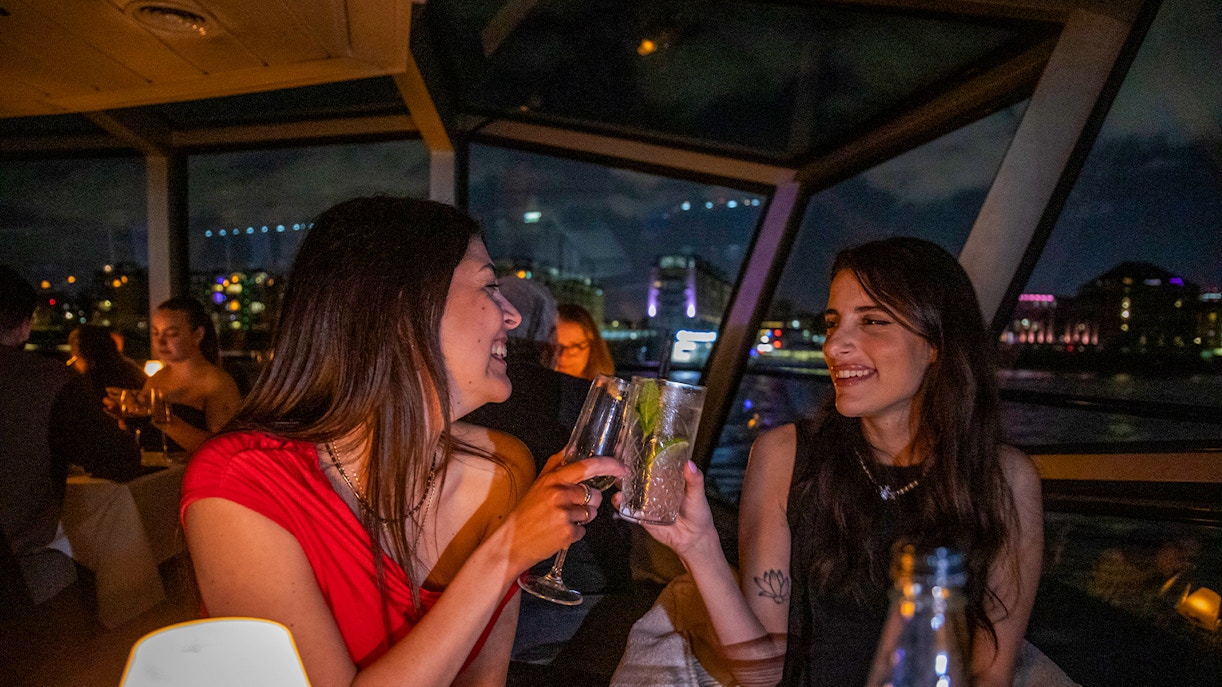 Guests enjoying evening drinks on the Thames River Cruise with London skyline in the background.