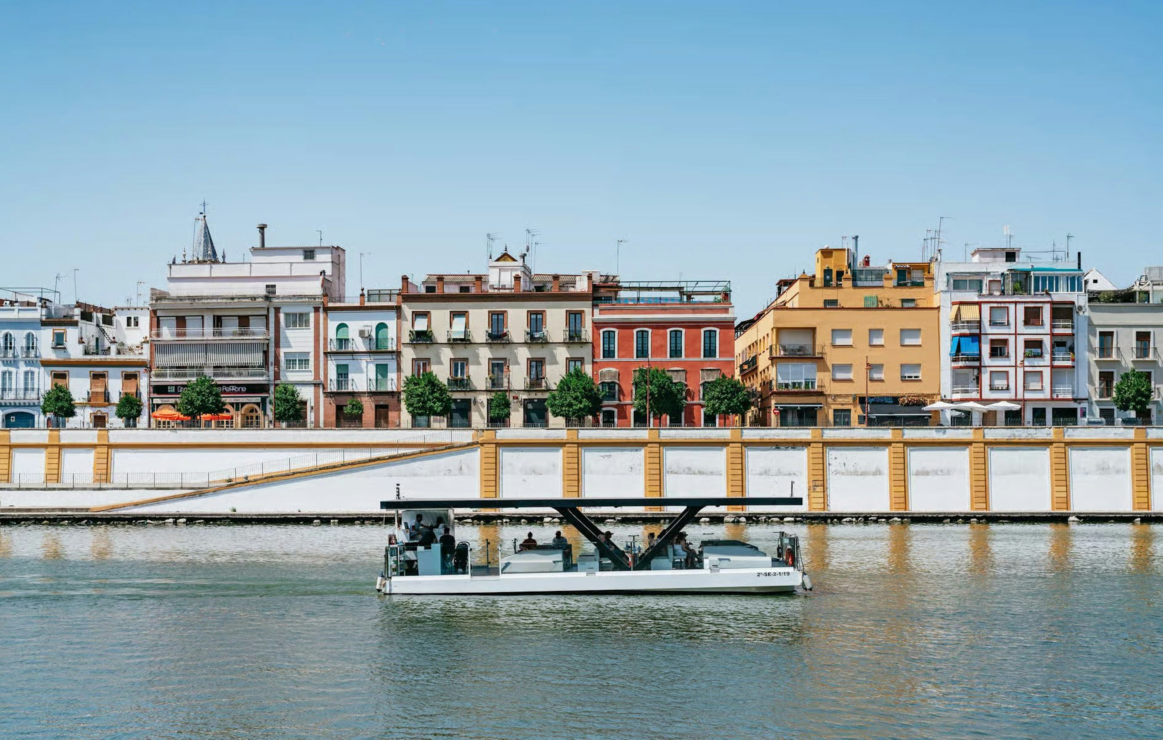 Cruise boat on Guadalquivir River with colorful buildings in Seville, Spain.