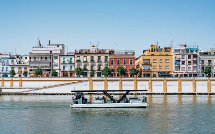 Cruise boat on Guadalquivir River with colorful buildings in Seville, Spain.
