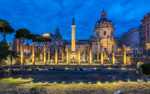 Trajan's Column illuminated at night in Rome, surrounded by ancient ruins and historic buildings.