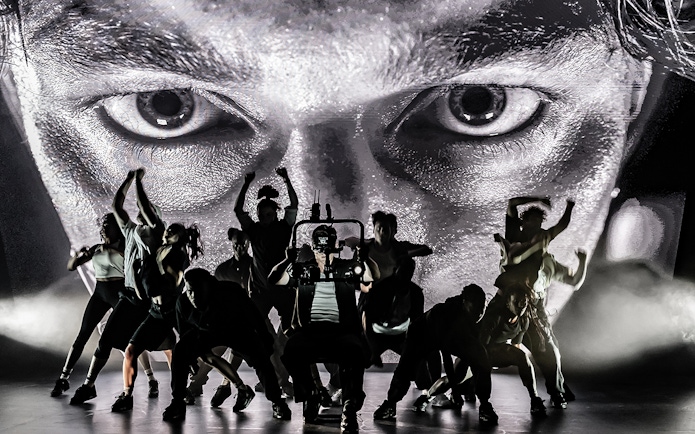 Dancers performing on stage with a large face projected behind them, Sunset Boulevard.