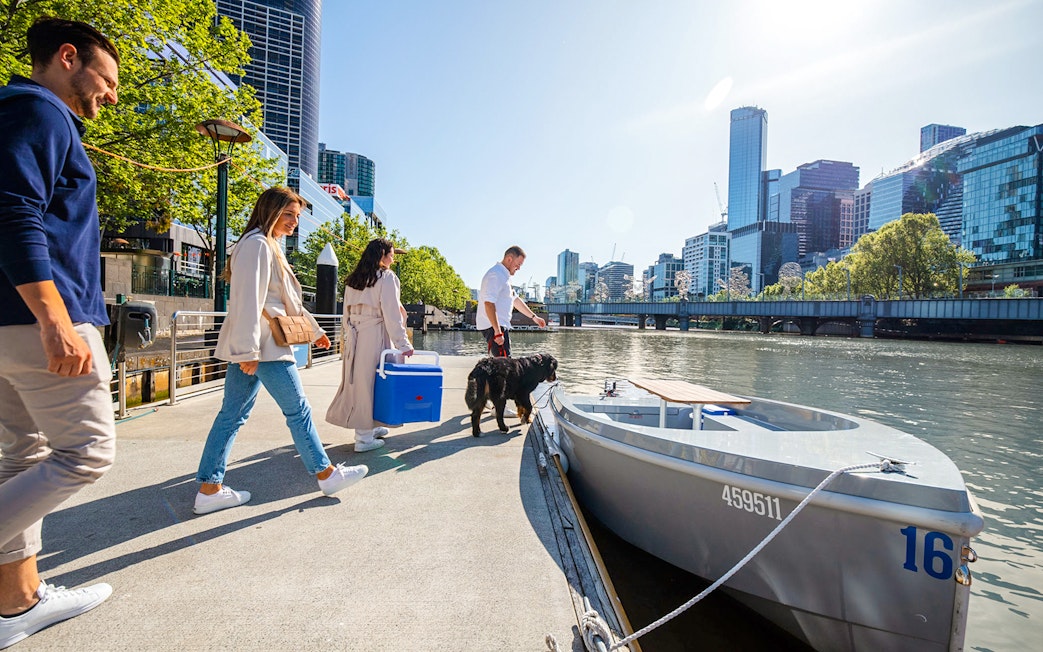 People boarding a self-drive picnic cruise on Yarra River with city skyline in background.