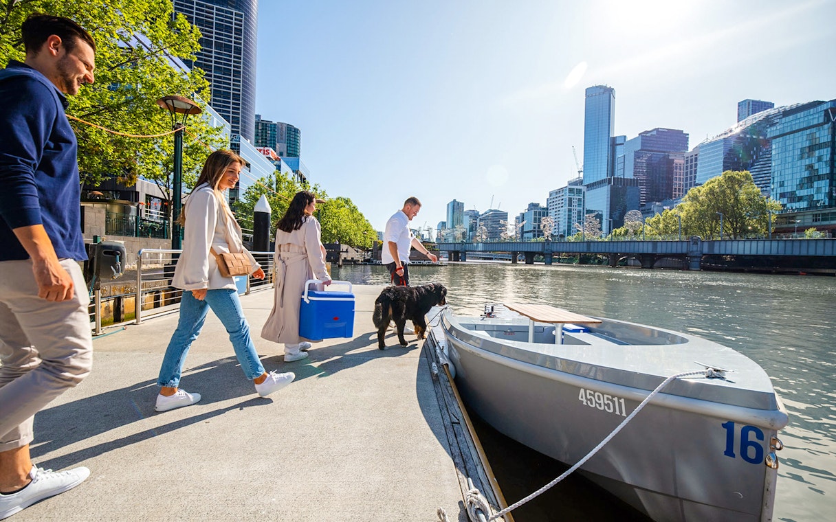 People boarding a self-drive picnic cruise on Yarra River with city skyline in background.