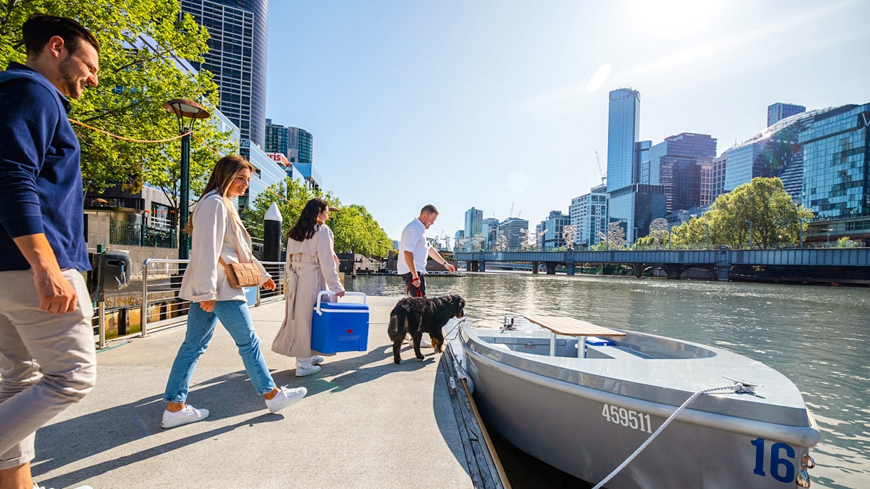People boarding a self-drive picnic cruise on Yarra River with city skyline in background.