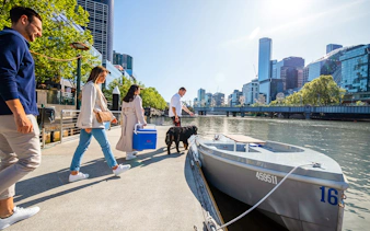 People boarding a self-drive picnic cruise on Yarra River with city skyline in background.