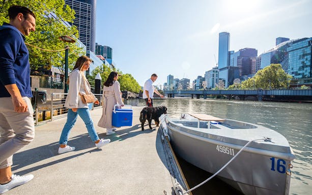 People boarding a self-drive picnic cruise on Yarra River with city skyline in background.
